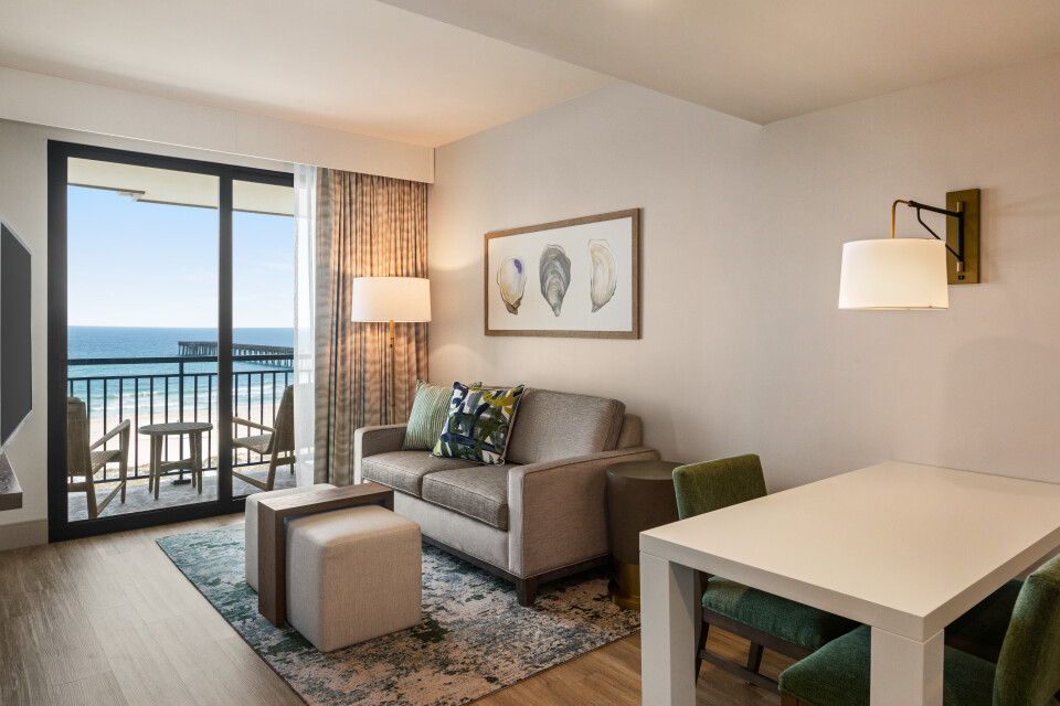 Hotel living room with ocean-view balcony, beige sofa, and two ottomans facing a coffee table and rug.