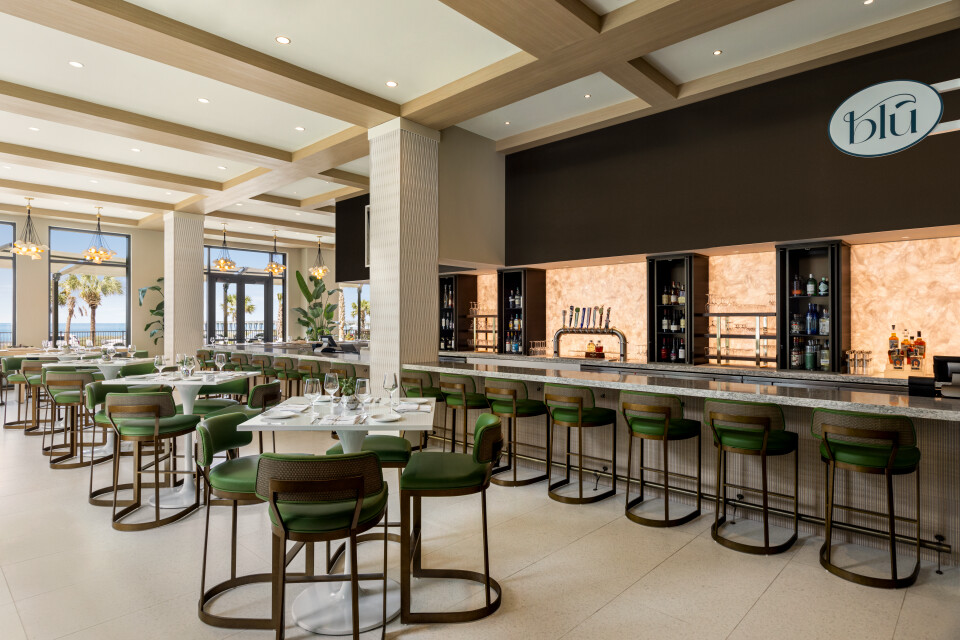 Bright beachfront restaurant interior with a long marble bar, green bar stools, and white tabletops near large windows overlooking the ocean.
